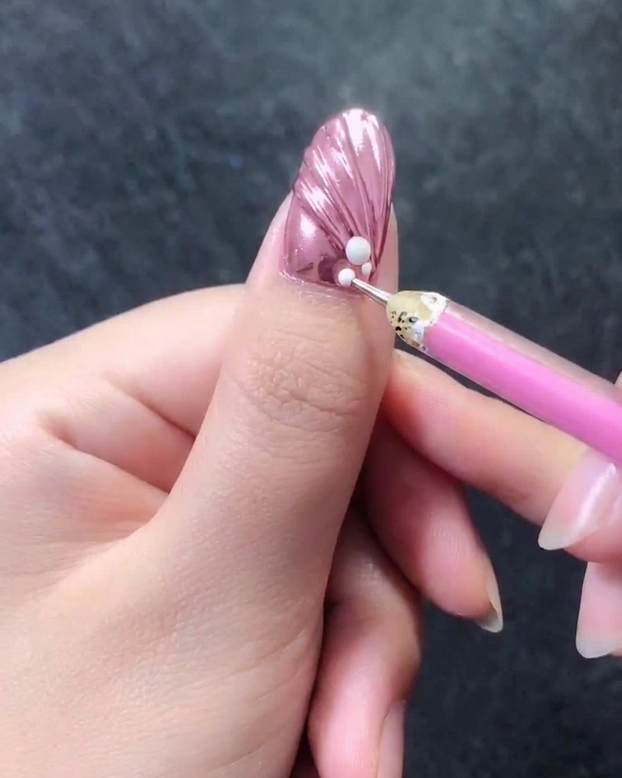 Gros plan dun ongle texturé rose métallique avec un stylo à cire ajoutant de minuscules perles blanches directement à côté dune perle plus grande sur la cuticule.