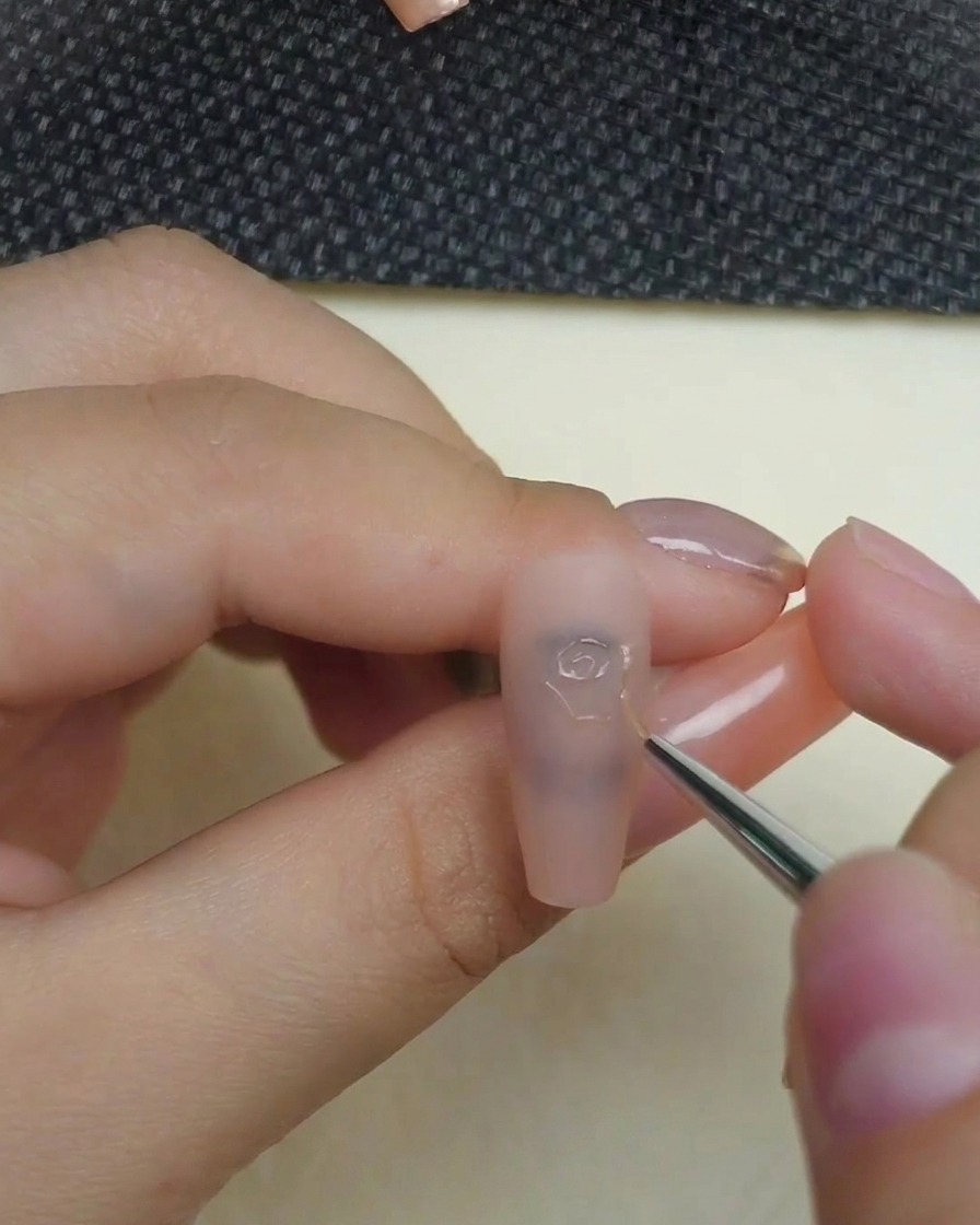 Gros plan dune main utilisant un pinceau liner fin pour appliquer du gel de sculpture 3D transparent, dessinant le tourbillon central précis dune rose sur un ongle nude mat.
