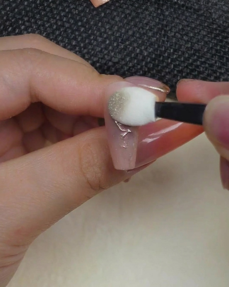 Un applicateur éponge type ombre à paupières pressant de la poudre chrome métallique sur un design de rose en relief sur un ongle.