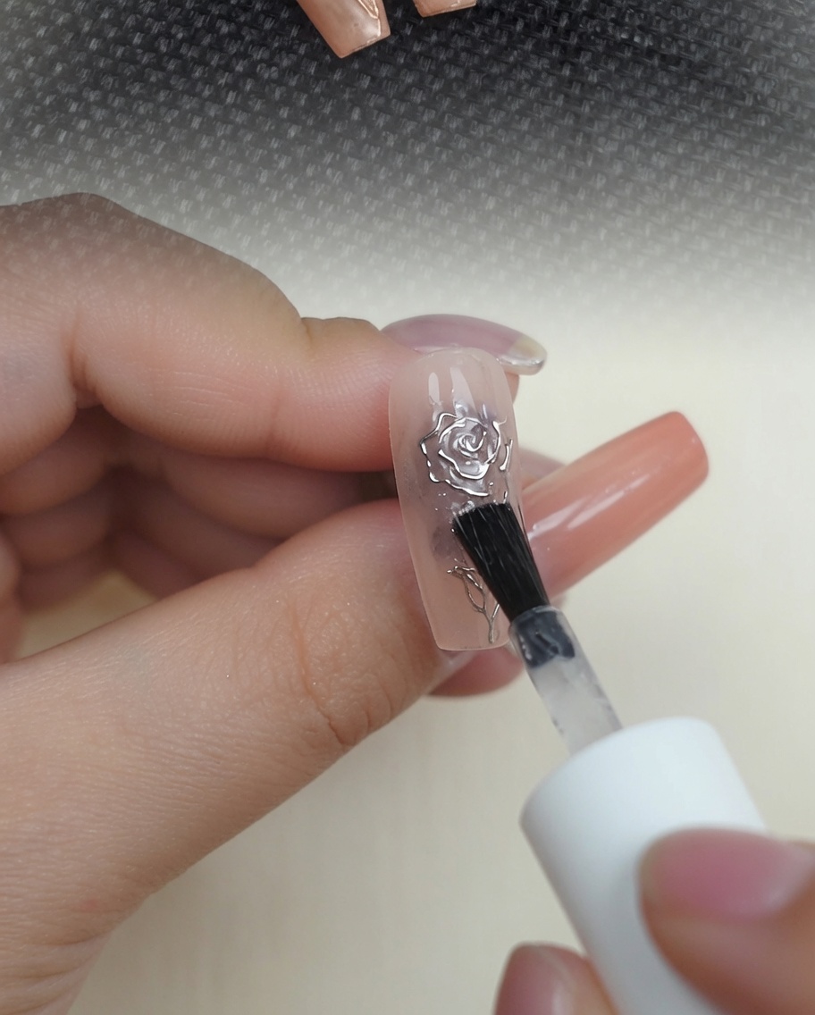 Un pinceau appliquant une couche de finition brillante et transparente sur un design de rose 3D chrome métallique sur un ongle nude.