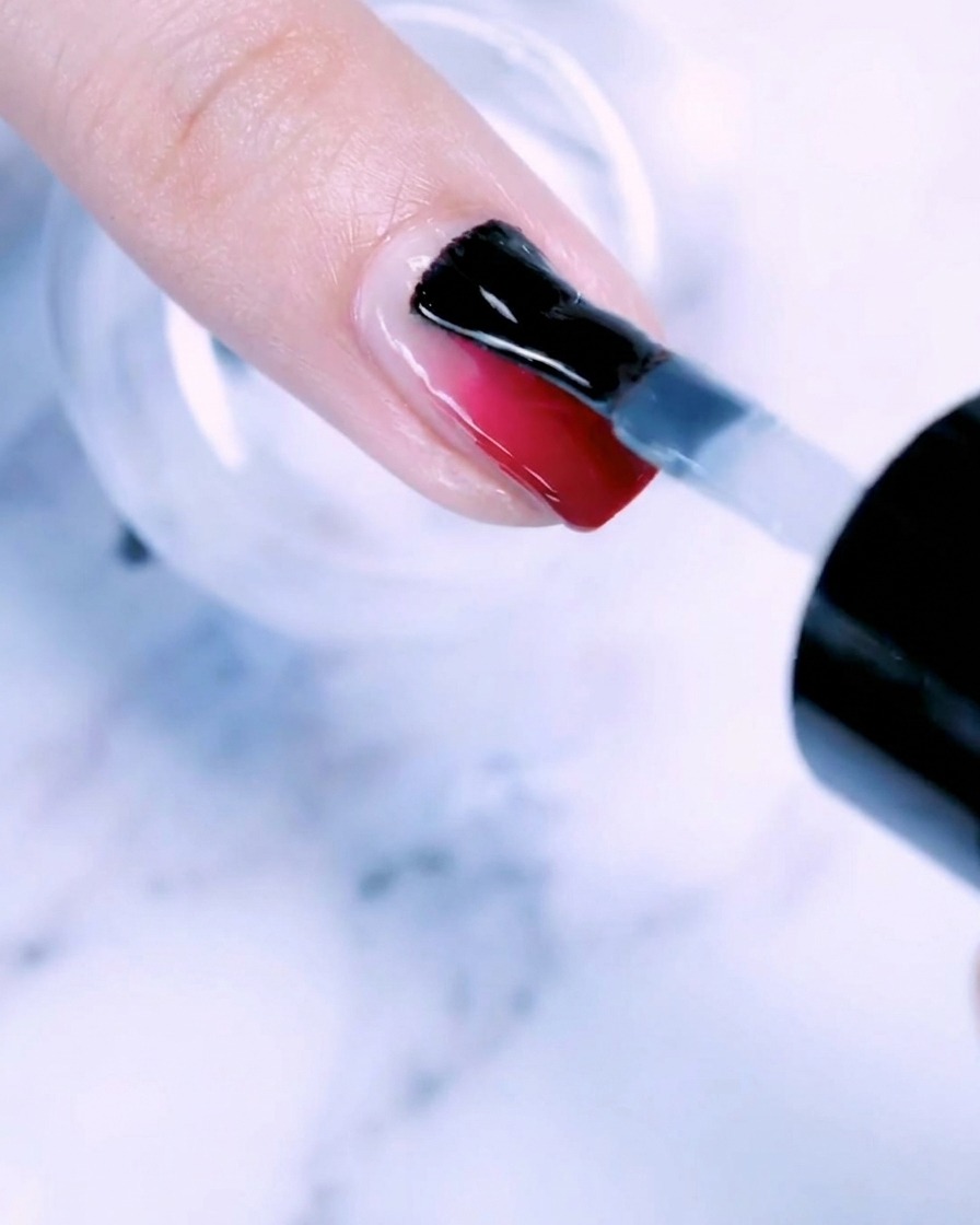 Un pinceau appliquant une couche de base transparente sur un ongle avec un dégradé or rose.