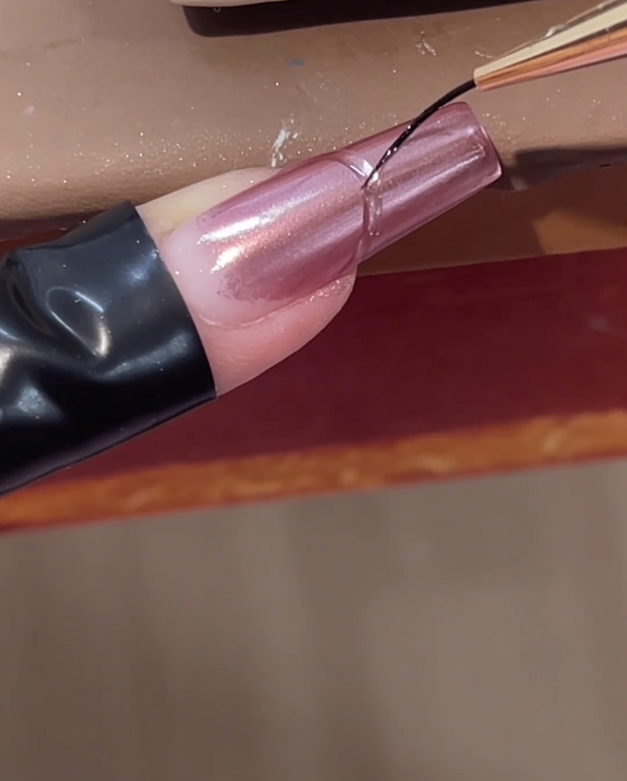 Un pinceau liner fin ajoutant une ligne de gel de construction transparent courbée près de la cuticule dun ongle chromé rose.