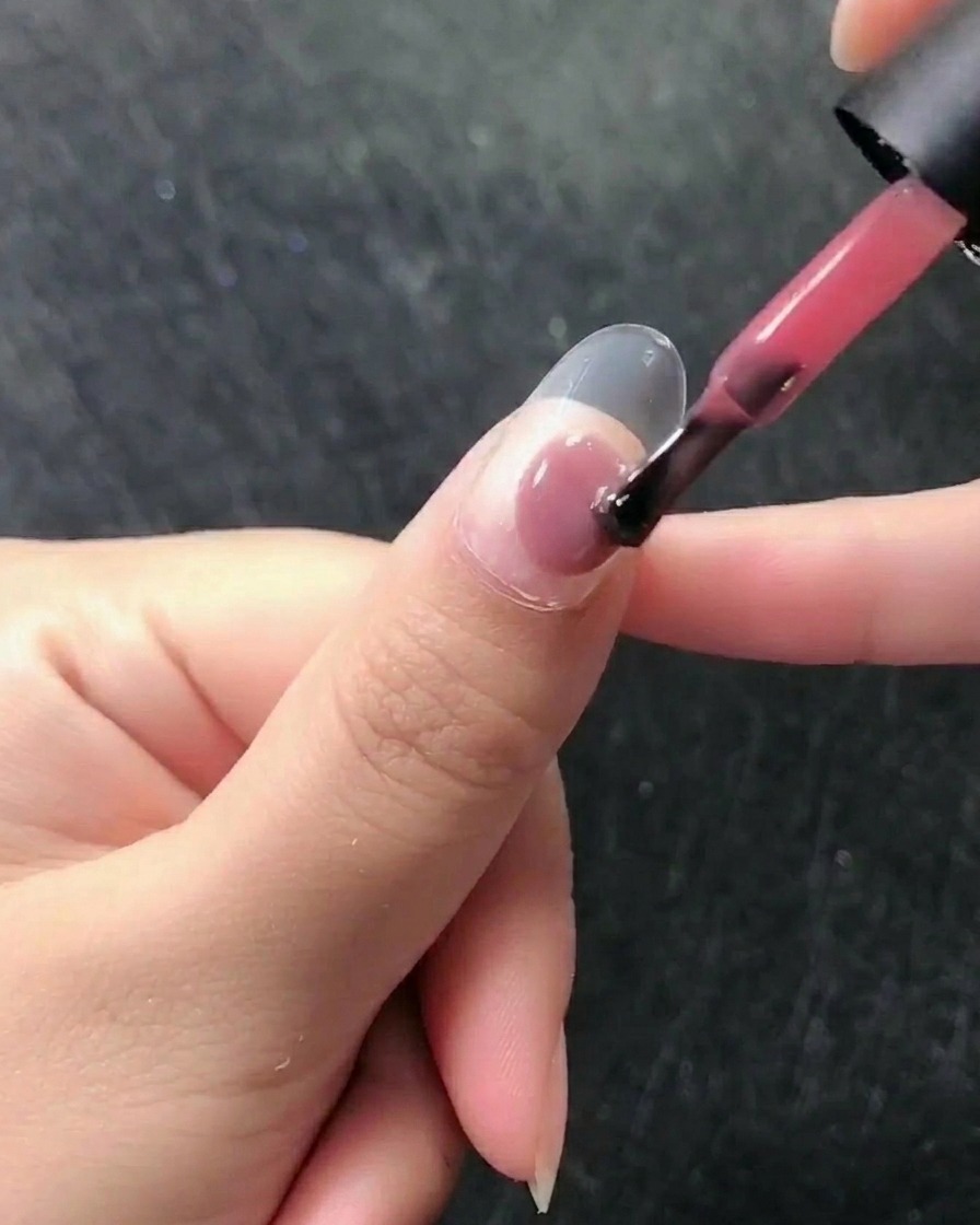 Gros plan dun pinceau appliquant un vernis gel rose translucide sur un ongle naturel préparé.