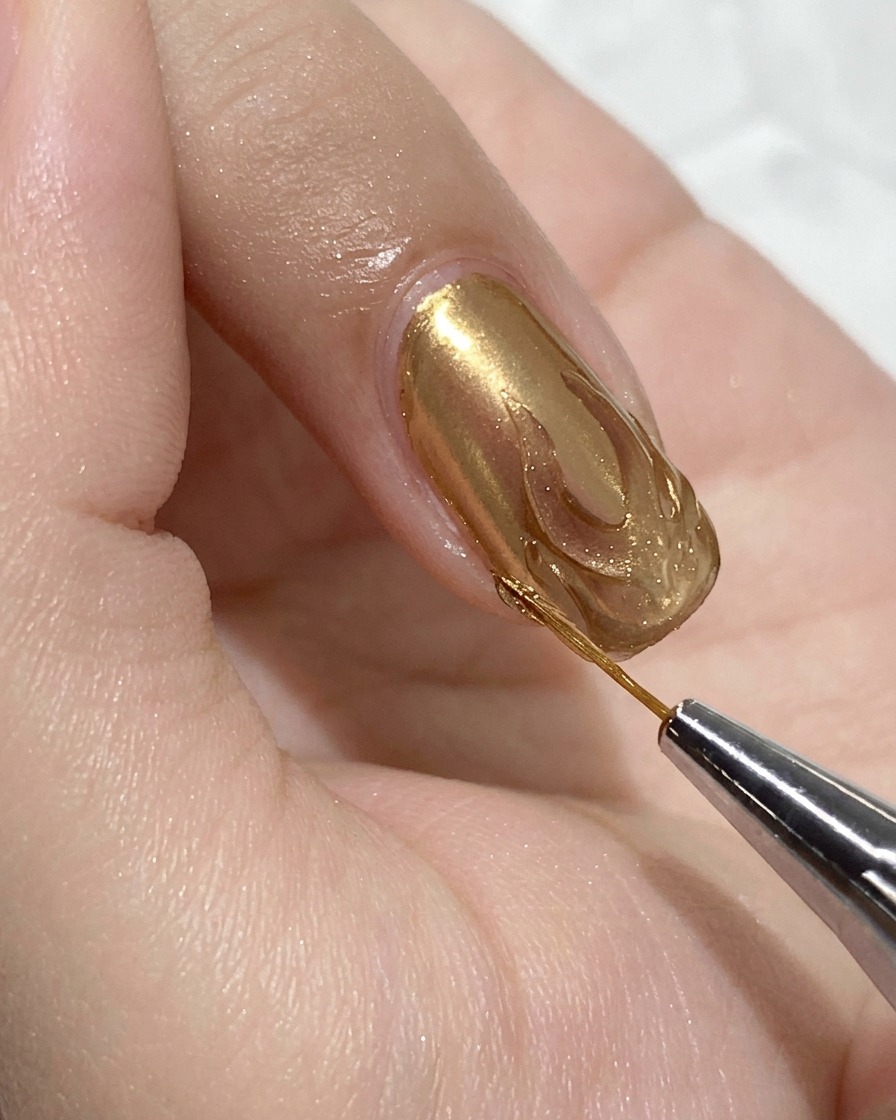 Gros plan sur un pinceau de détail fin appliquant un top coat gel de construction à lintérieur dun contour de flamme chromé doré sur un ongle rose.