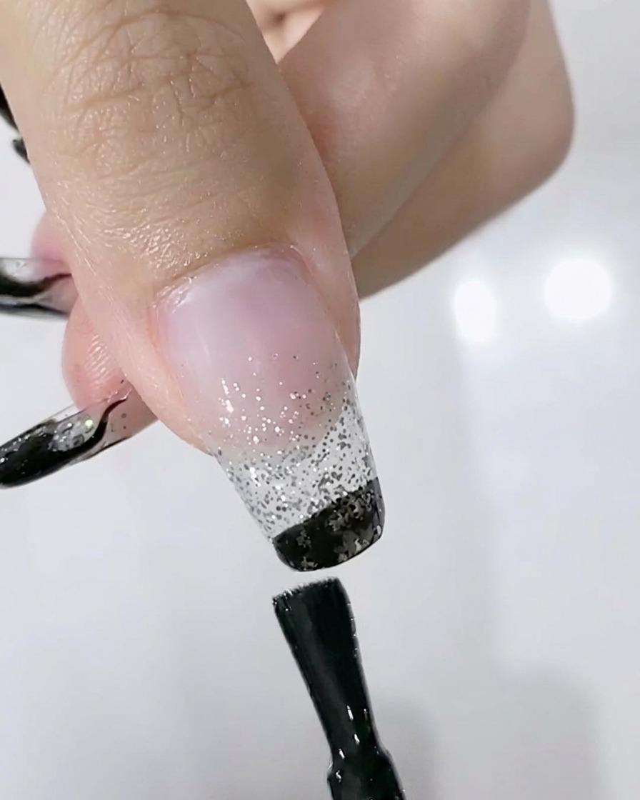 Un pinceau appliquant du vernis gel noir sur le bord libre dun ongle par-dessus un dégradé pailleté.