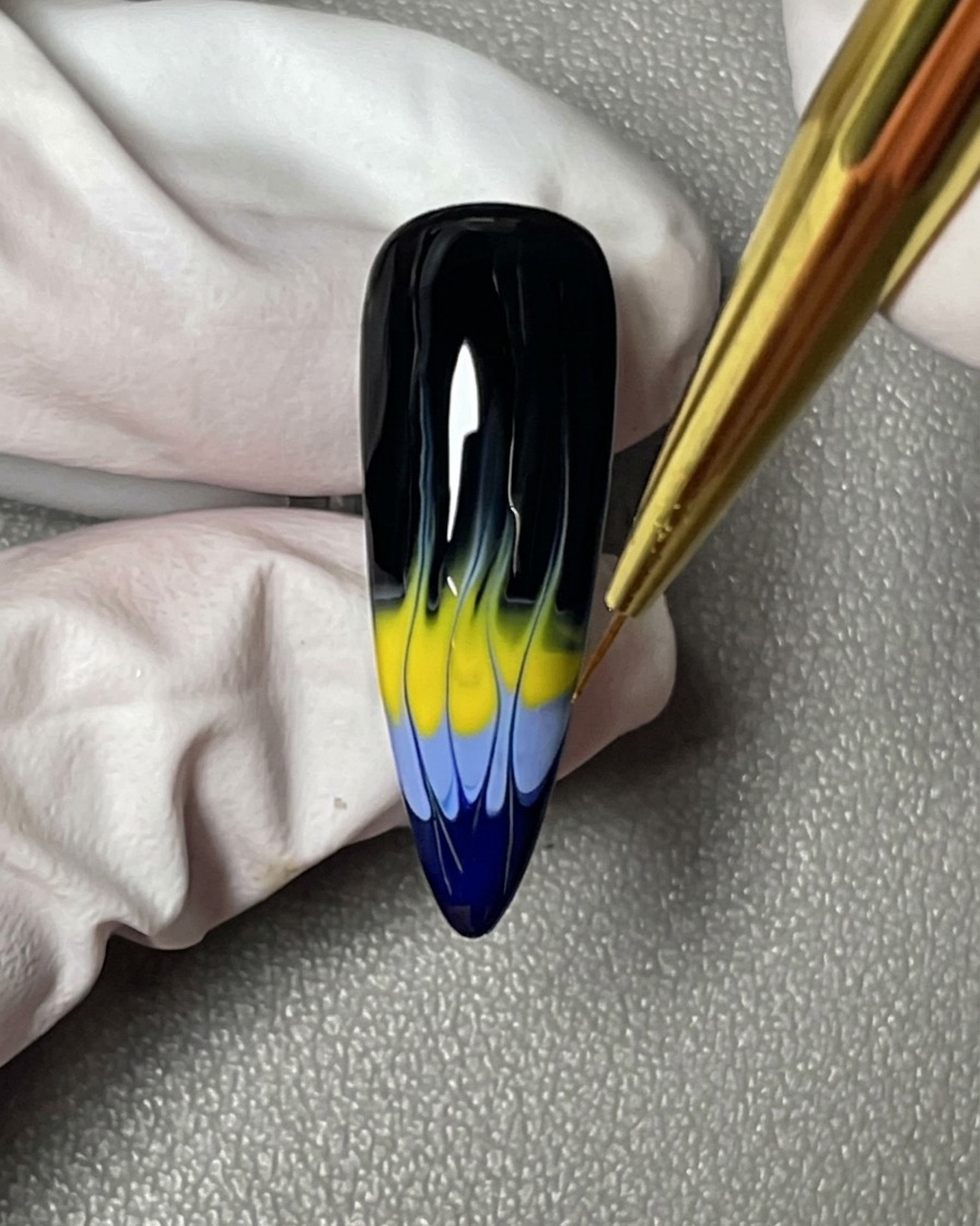 Un gros plan montrant un pinceau liner perfectionnant les pointes acérées du design de flammes bleu et jaune sur un ongle noir.