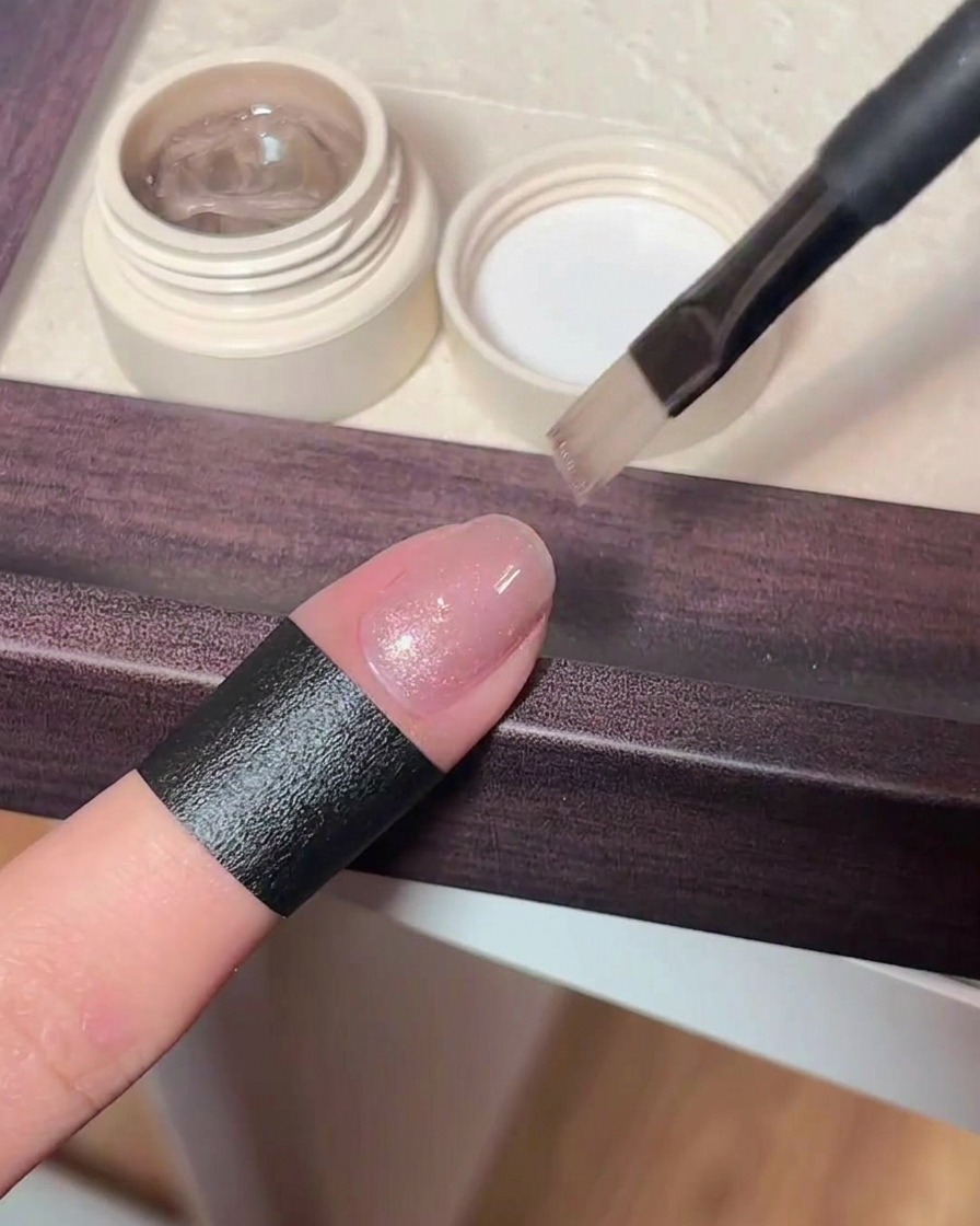 Gros plan sur un pinceau appliquant un gel cat-eye rose scintillant et translucide sur un ongle naturel.