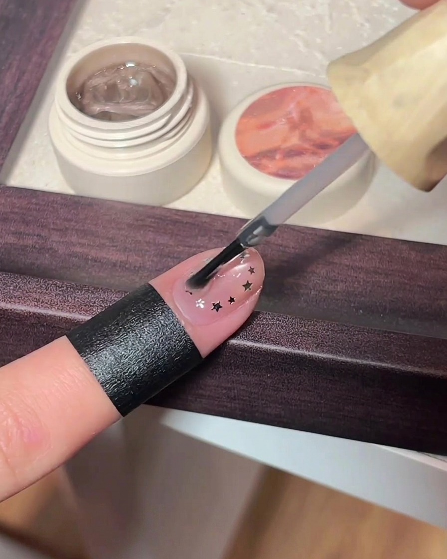 Un pinceau appliquant une couche épaisse de top coat transparent sur un ongle rose cat-eye décoré de petites étoiles.