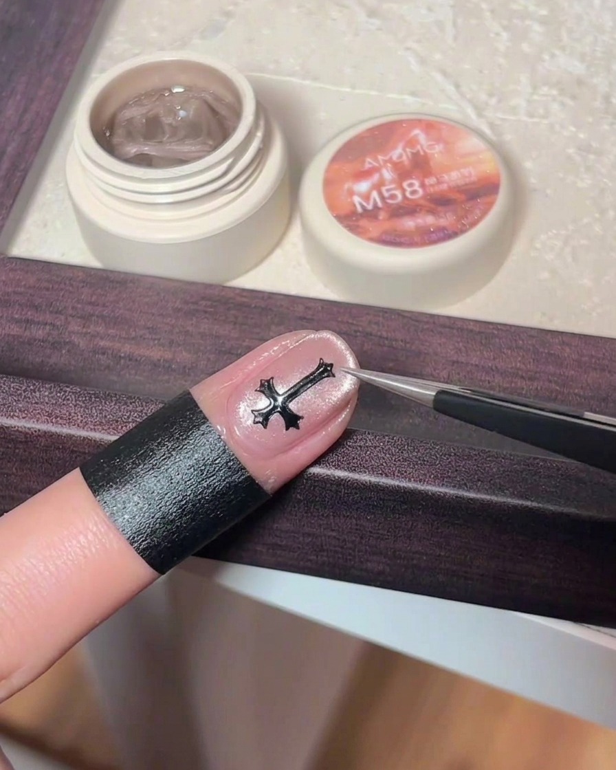 Une pince déposant une petite croix métallique noire au centre dun ongle en gel rose cat-eye.