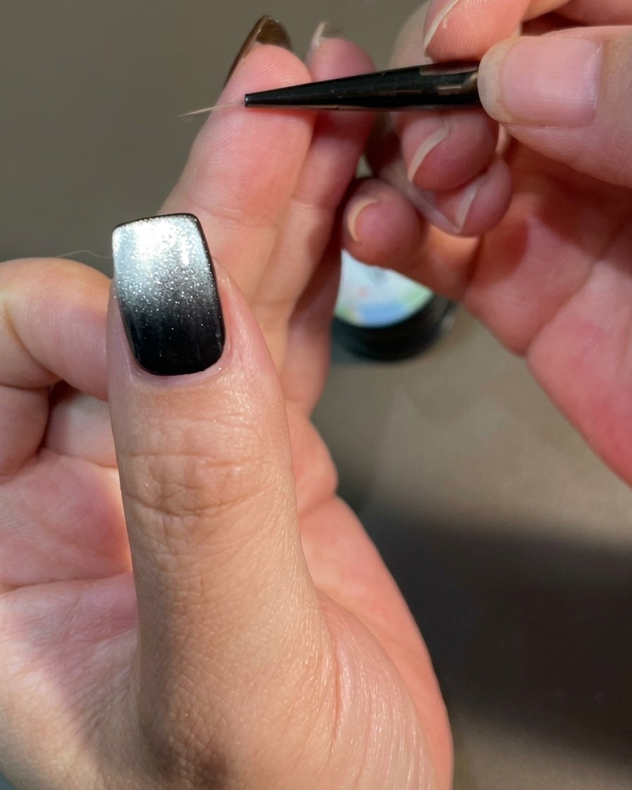 Vue rapprochée dun ongle décoré avec un pinceau fin créant des lignes courbes en forme de flamme sur une base de gel magnétique noire.