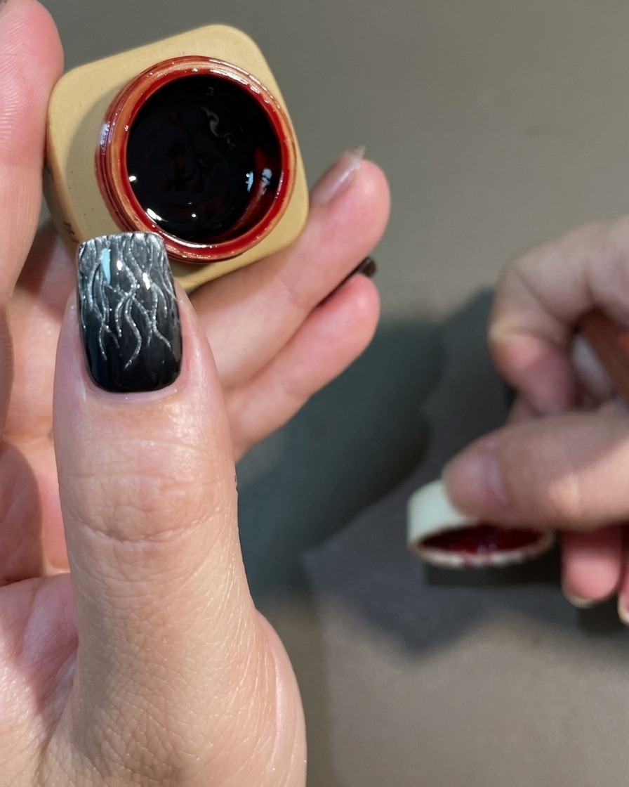 Un pinceau de nail art appliquant un gel rouge foncé transparent sur un motif de flamme cat-eye noir et blanc catalysé.