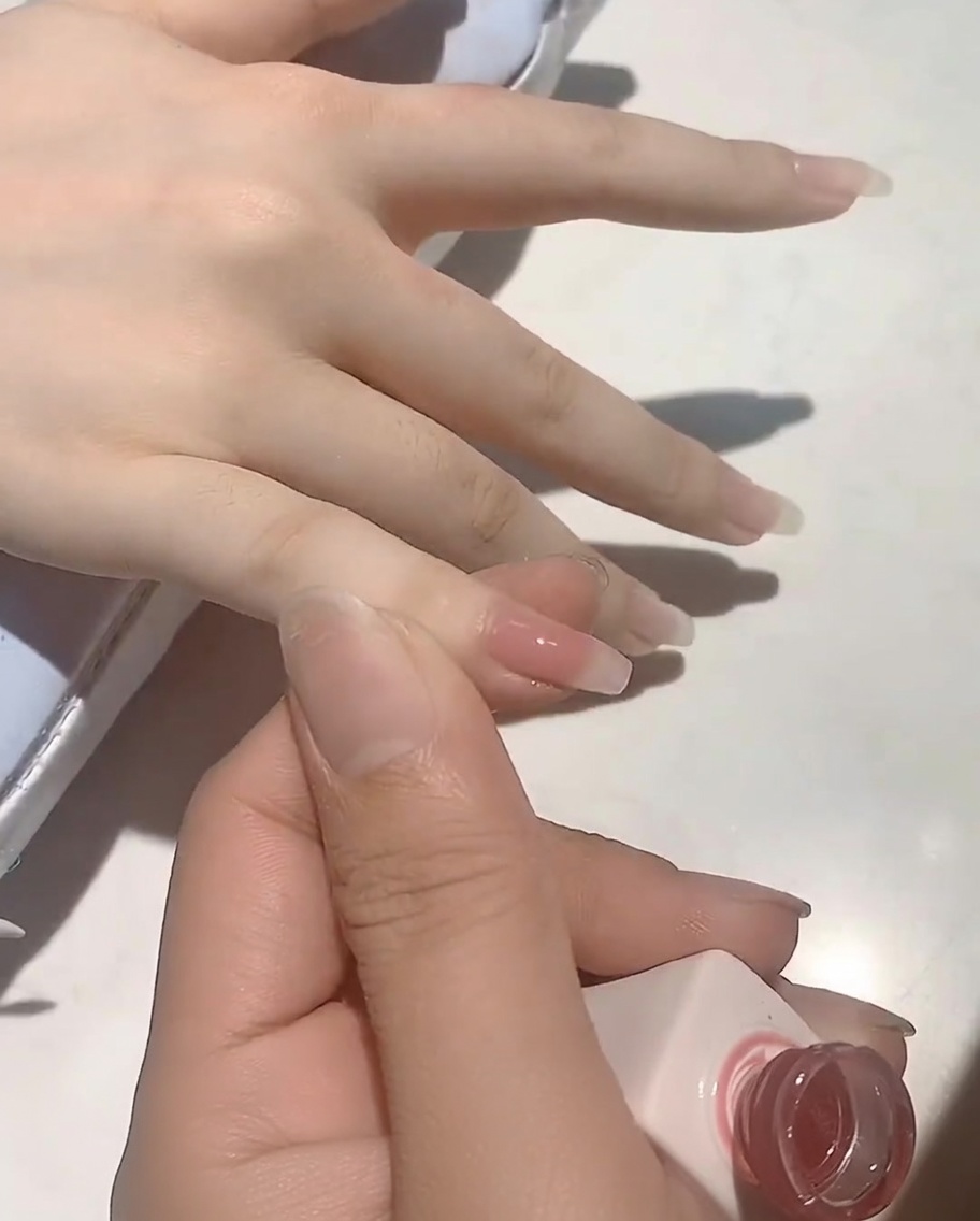 Un pinceau appliquant un gel de base rose nude doux sur un ongle naturel.