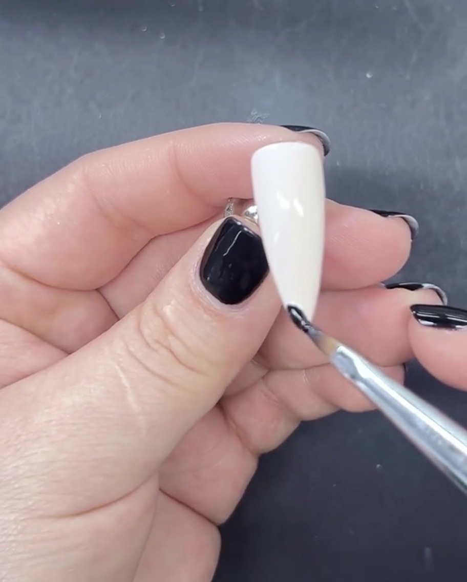 Un pinceau liner fin appliquant une petite ligne précise de vernis gel noir sur le bout dun ongle peint en blanc.