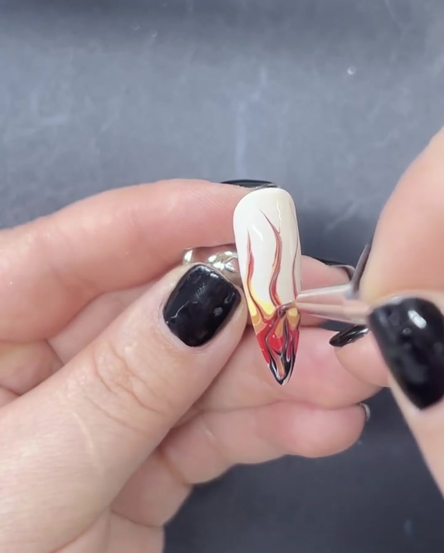 Gros plan dun pinceau liner étirant du vernis gel rouge et noir vers le haut sur une base blanche pour créer des formes de flammes.