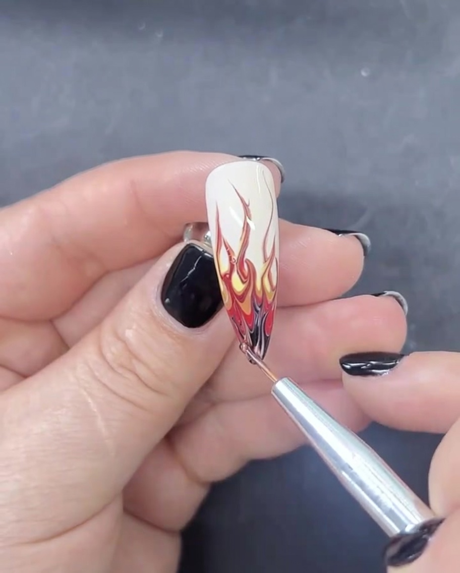 Un pinceau liner appliquant une minuscule quantité de vernis gel noir sur le bout dun ongle en amande pour finir un motif de flamme.