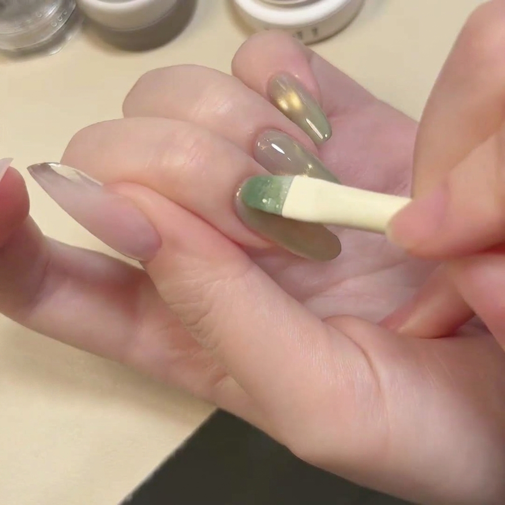 Un pinceau appliquant une couche de vernis gel vert lagon translucide et chatoyant sur longle.