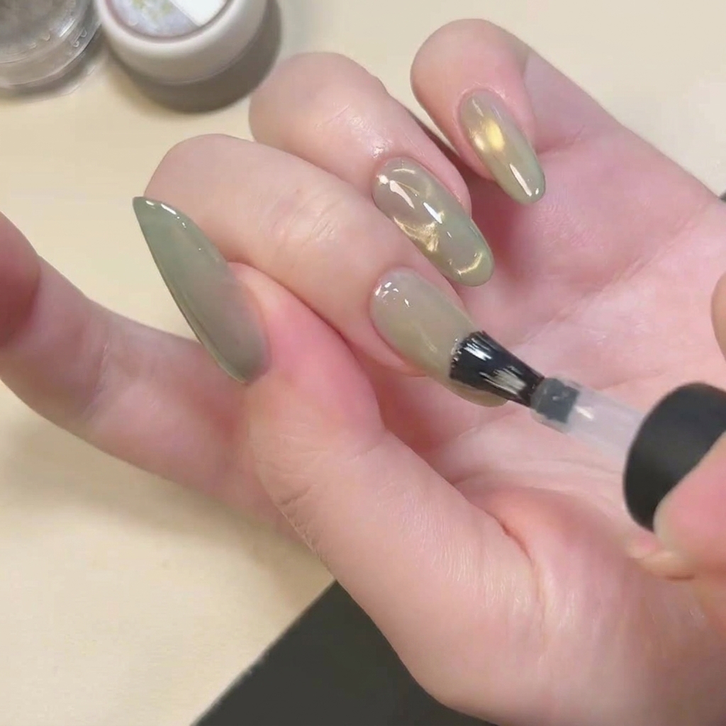 Un pinceau appliquant un top coat transparent à finition mate sur un ongle cat eye vert.