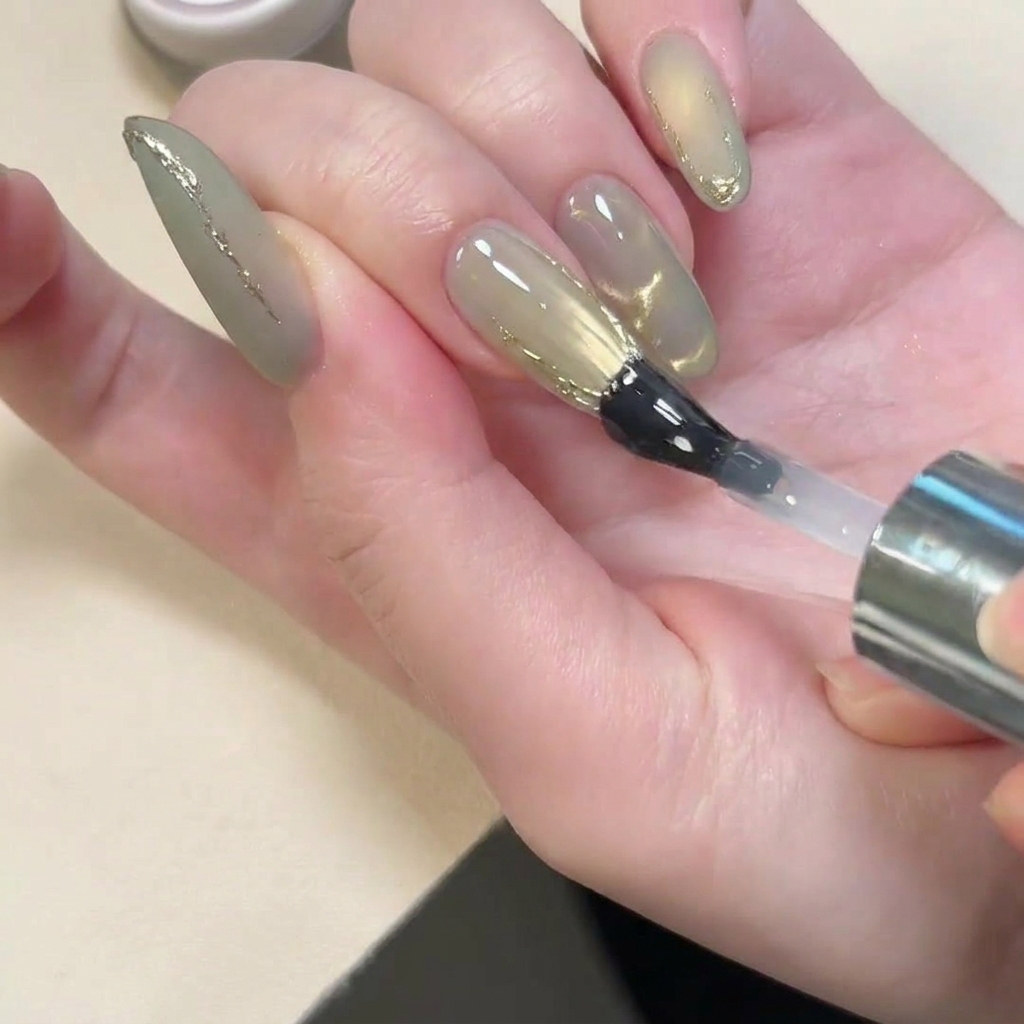 Un pinceau appliquant un top coat brillant sur un ongle au fini mat avec des accents 3D dorés.