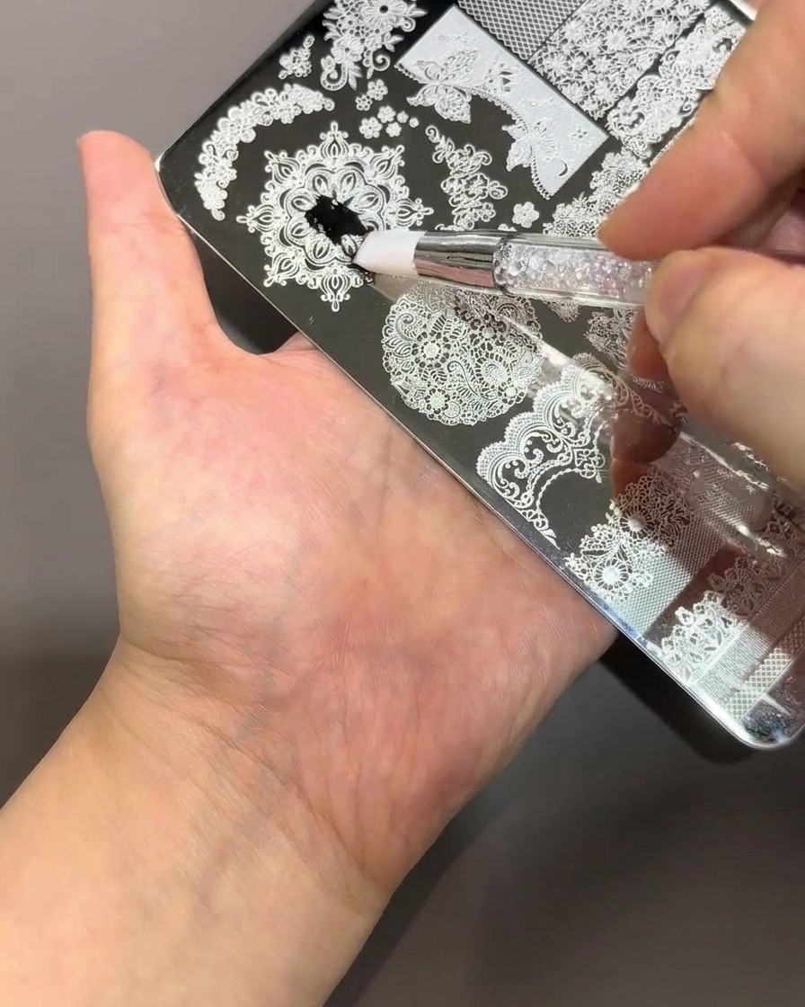 Un pinceau applique un vernis noir épais sur une plaque de stamping en métal avec des motifs de dentelle et floraux.