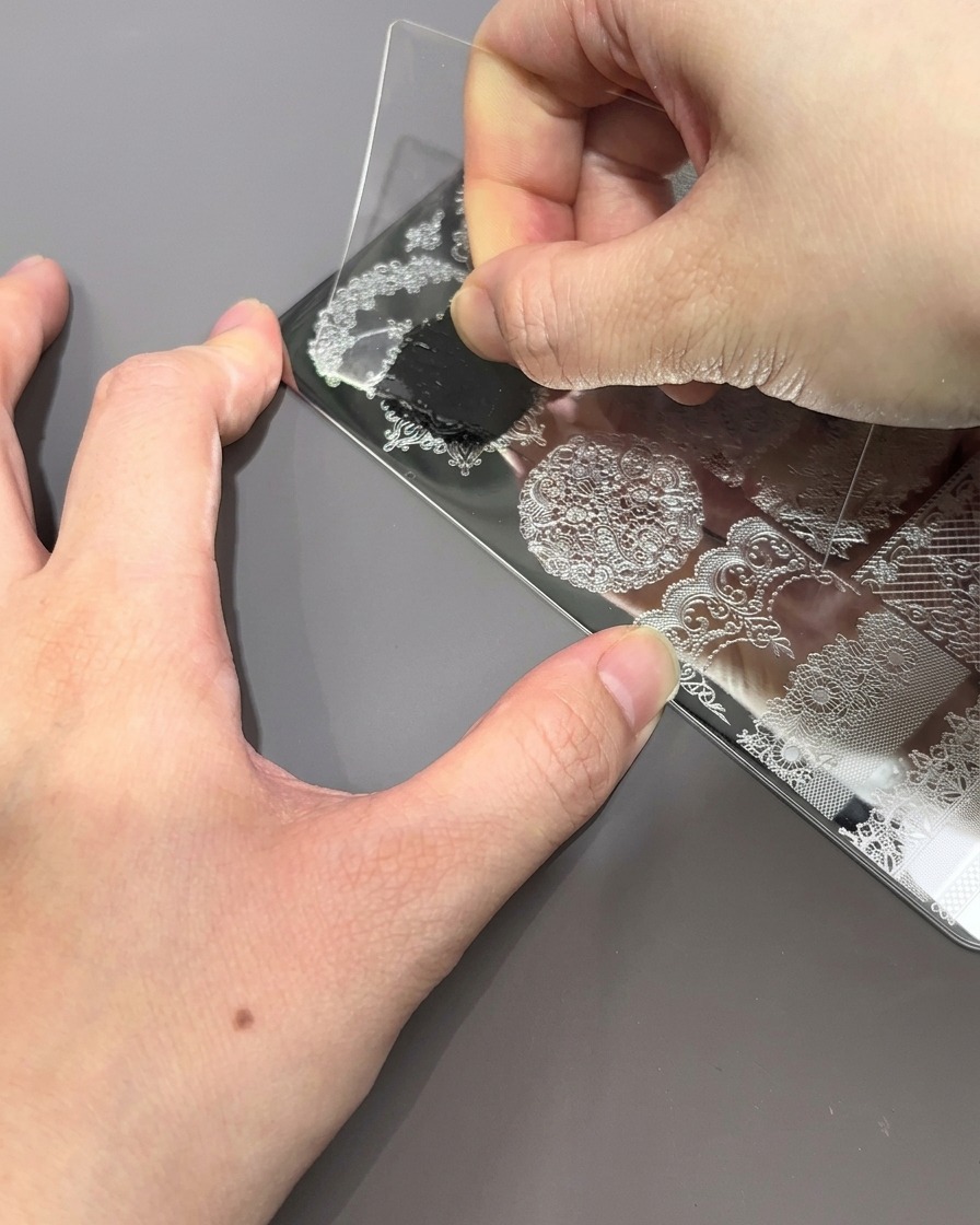 Une carte raclette en plastique transparent est tirée sur une plaque de stamping, poussant lexcès de vernis noir.