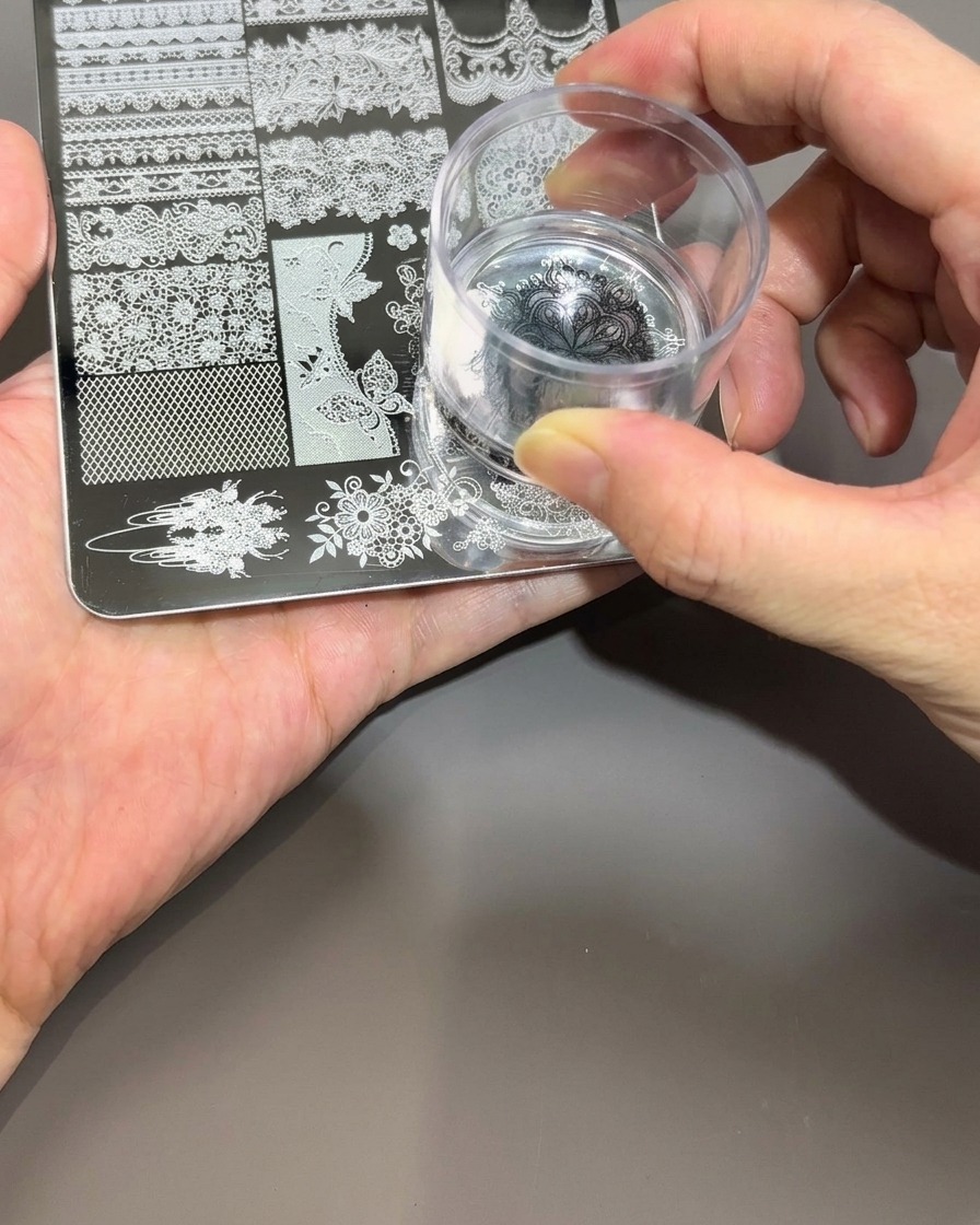 Une main tenant un tampon en silicone transparent, pressant doucement sur le motif noir dune plaque de stamping.