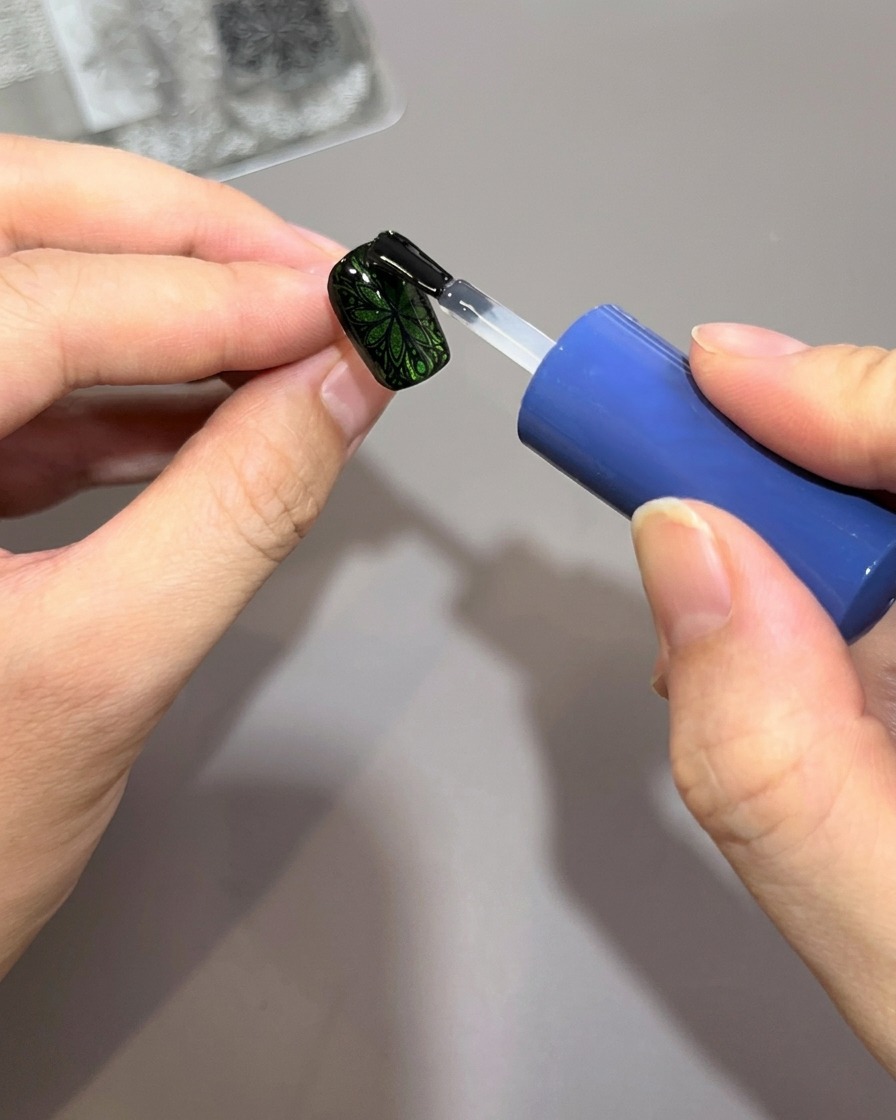 Un pinceau à vernis applique une couche épaisse de top coat brillant sur un ongle vert décoré dun motif noir.