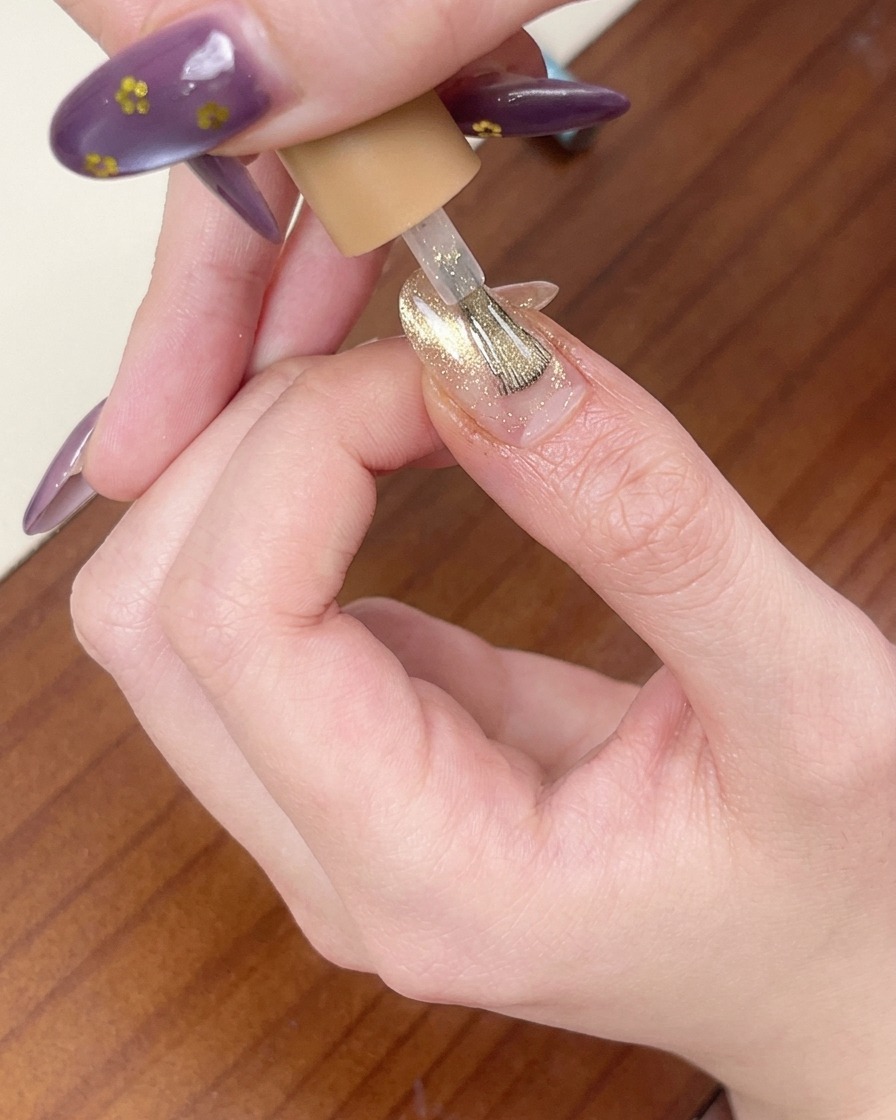 Un pinceau appliquant une couche de vernis gel œil-de-chat rose doré scintillant sur longle.