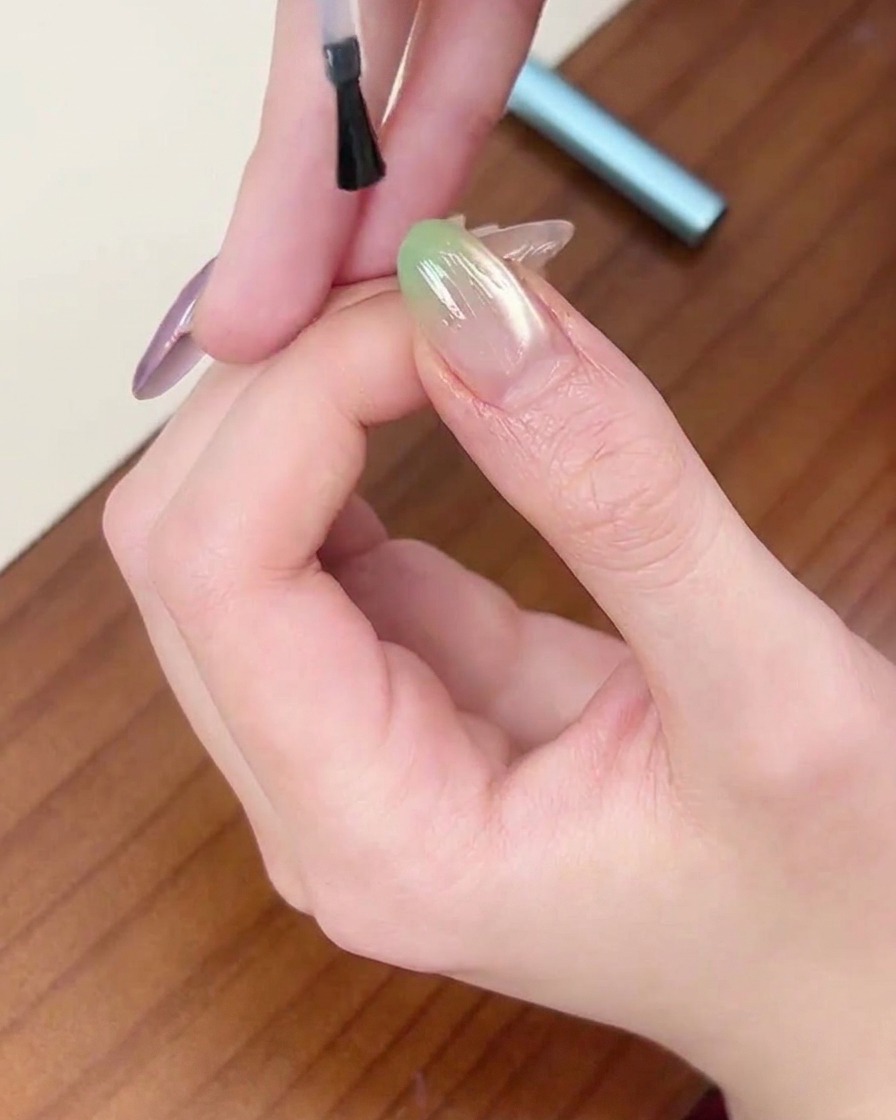 Un pinceau appliquant une couche finale de top coat transparent sur un ongle en dégradé vert.