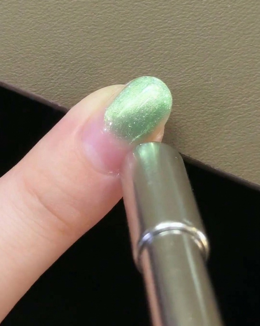 Un aimant métallique pour ongles tenu parallèlement au côté dun ongle recouvert de gel magnétique vert.