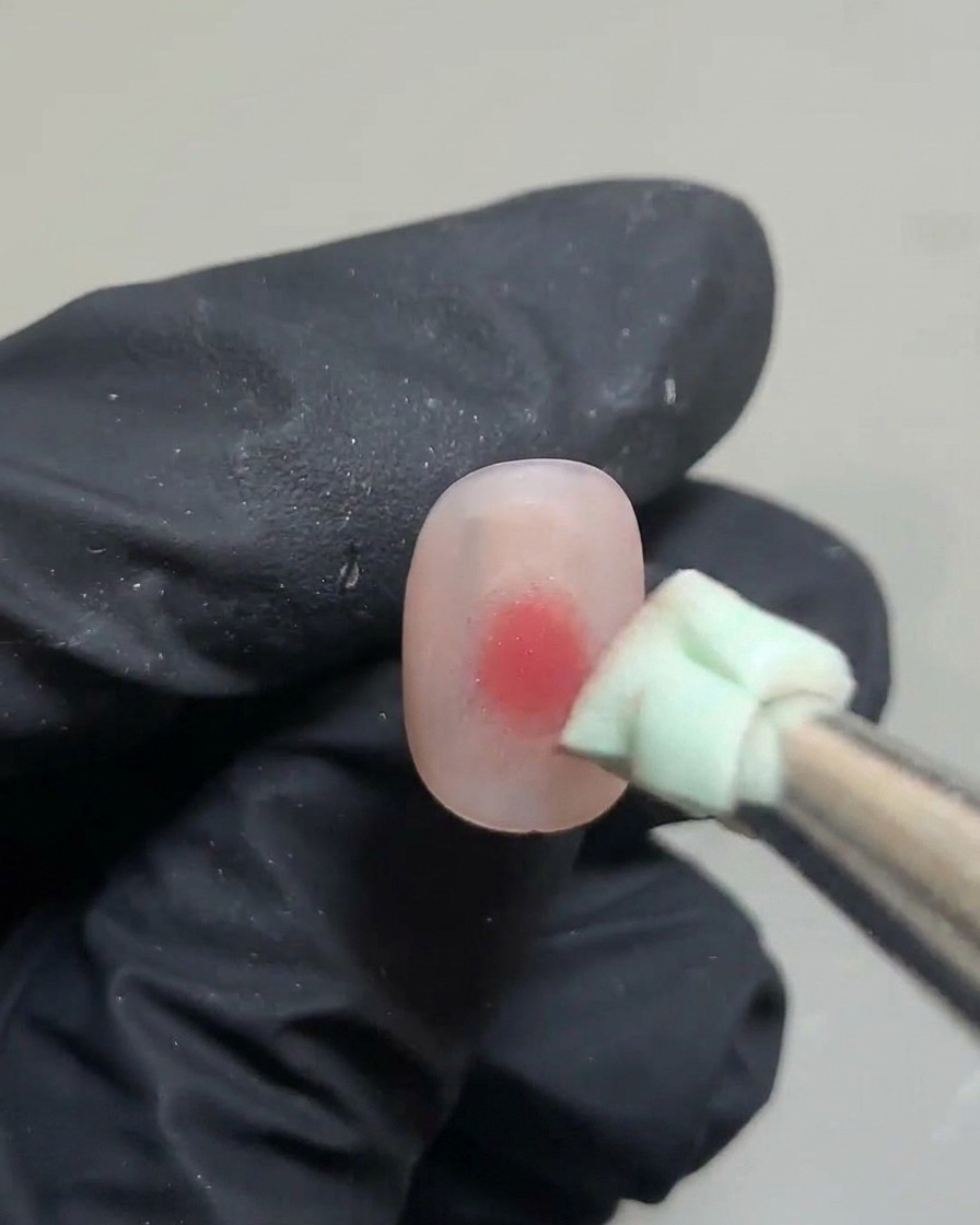 Une pince tenant un petit morceau déponge verte, tapotant un point de gel rose sur un ongle.