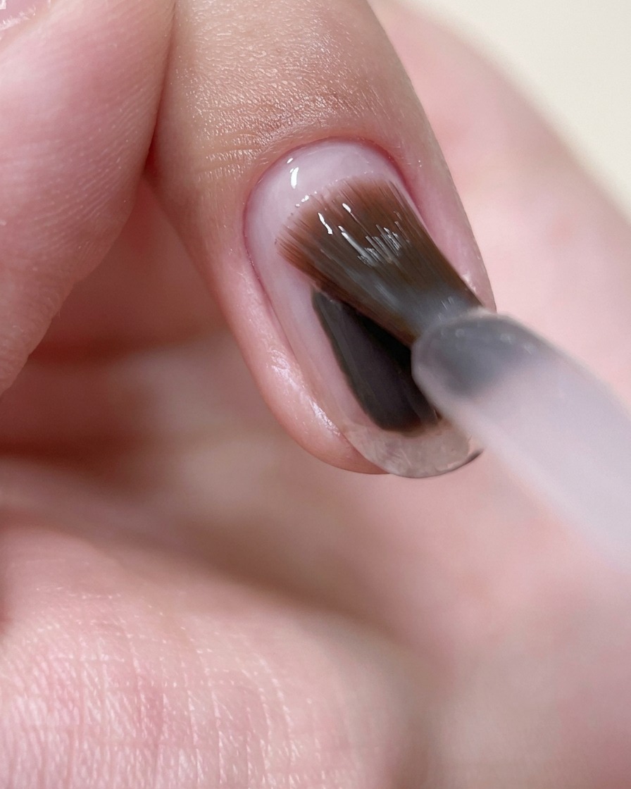 Vue rapprochée montrant un pinceau appliquant une fine couche de gel de construction rose transparent sur un ongle naturel.