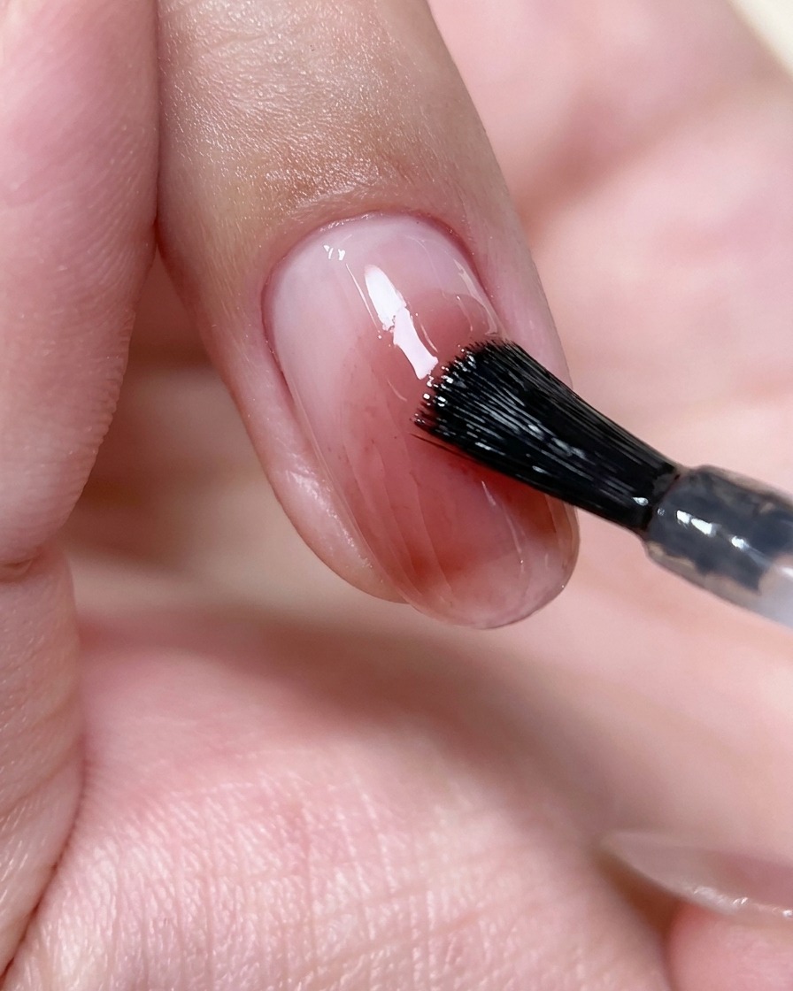 Un pinceau appliquant une fine couche transparente de top coat sur un ongle blush.