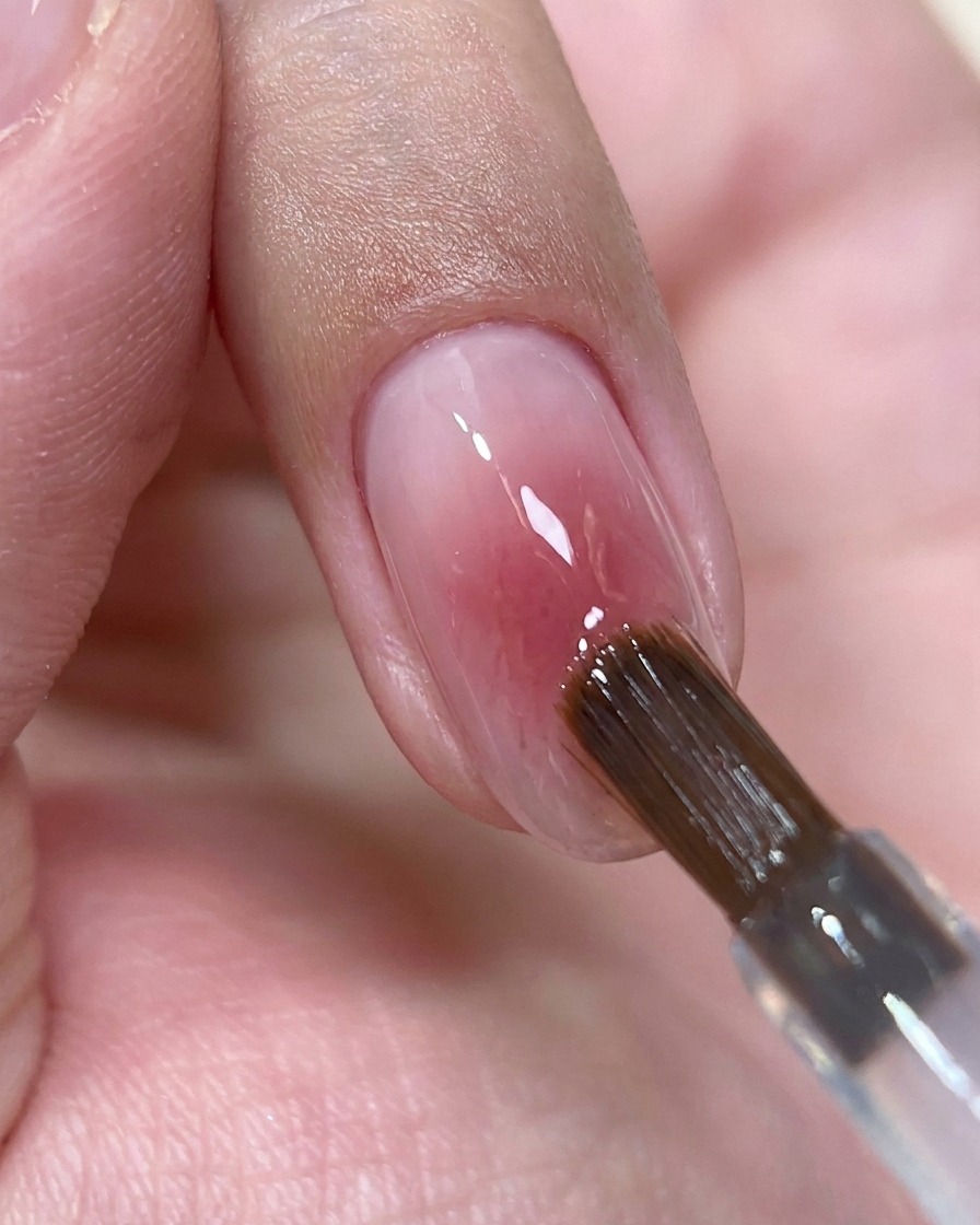 Un pinceau déposant une petite perle de top coat transparent au centre dun ongle blush brillant.