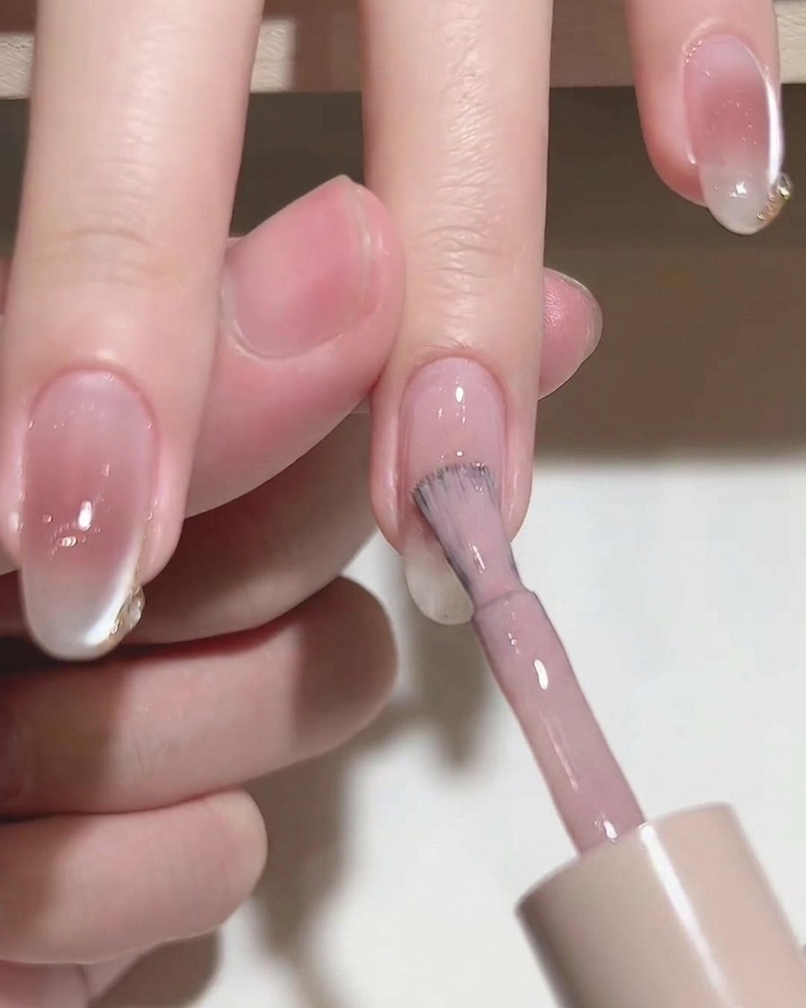 Gros plan dun pinceau appliquant un vernis gel rose translucide sur un ongle naturel.