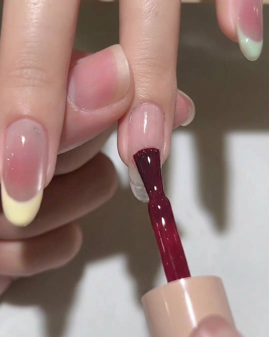 Un pinceau appliquant une couche de vernis gel rose translucide sur un ongle en forme damande.
