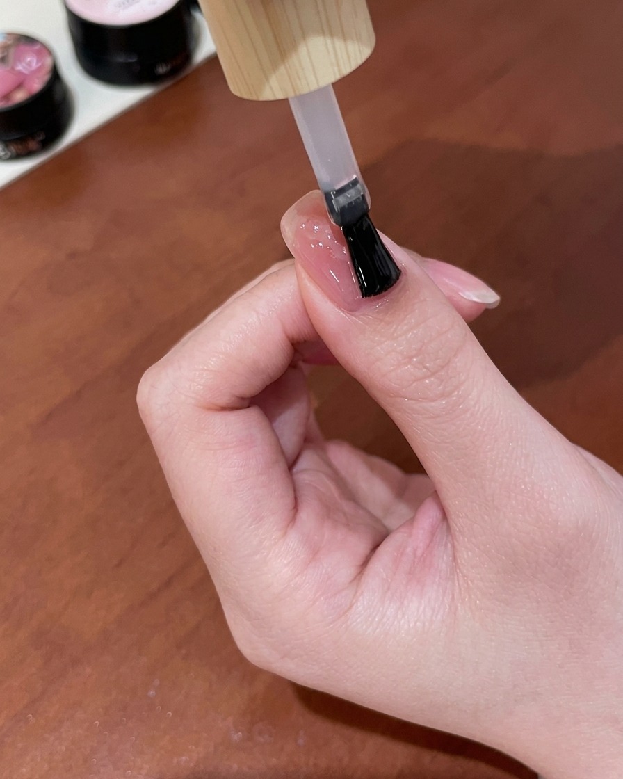 Un pinceau appliquant une couche de top coat gel transparent et brillant sur un ongle rose nud fini avec un design de papillon cat-eye en or.