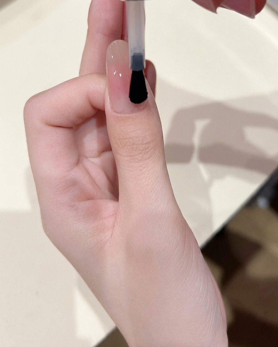 Un pinceau à ongles appliquant une fine couche de gel de base brillant et transparent sur un ongle ayant un centre blush doux.