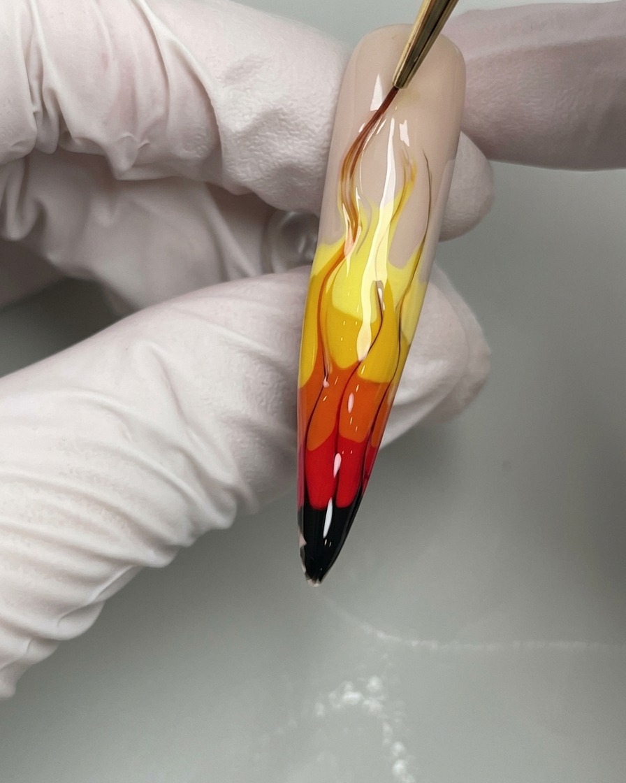 Gros plan dun pinceau à ongles appliquant de fines lignes de gel noir pour définir les bords dun nail art flammes jaune, orange et rouge.