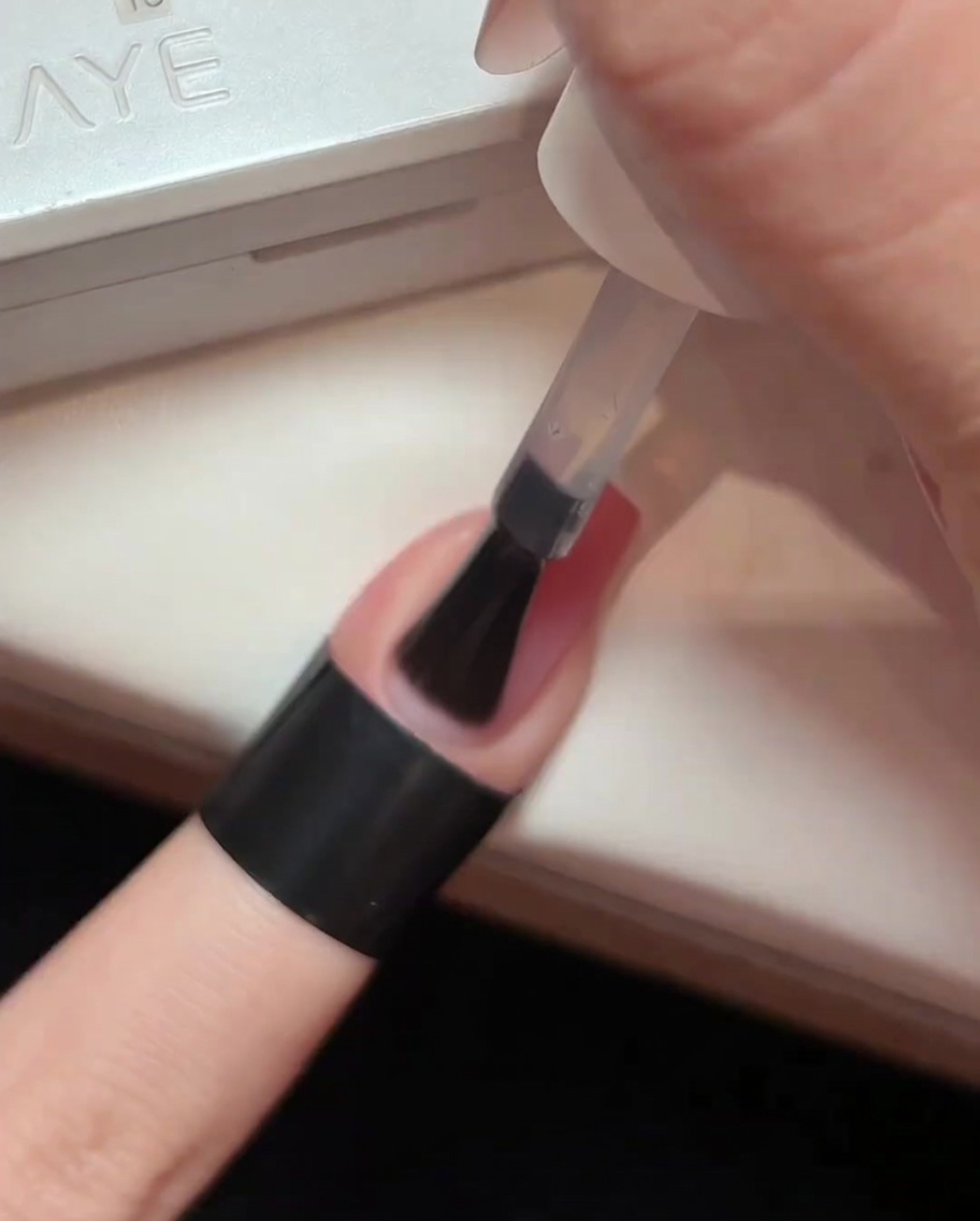 Un pinceau appliquant une couche de top coat brillant et transparent sur un ongle en dégradé rose clair.