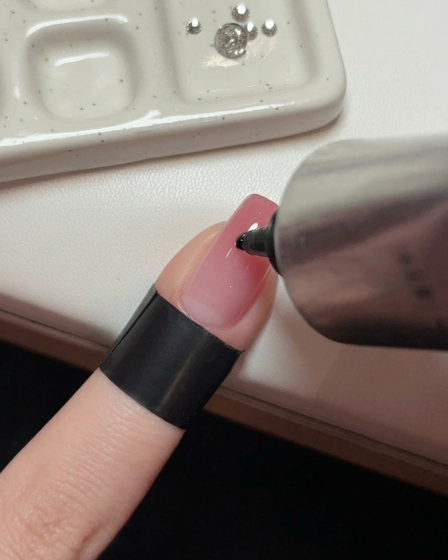 Une main tenant un tube métallique pressant une goutte transparente de gel adhésif au centre dun ongle rose brillant manucuré.
