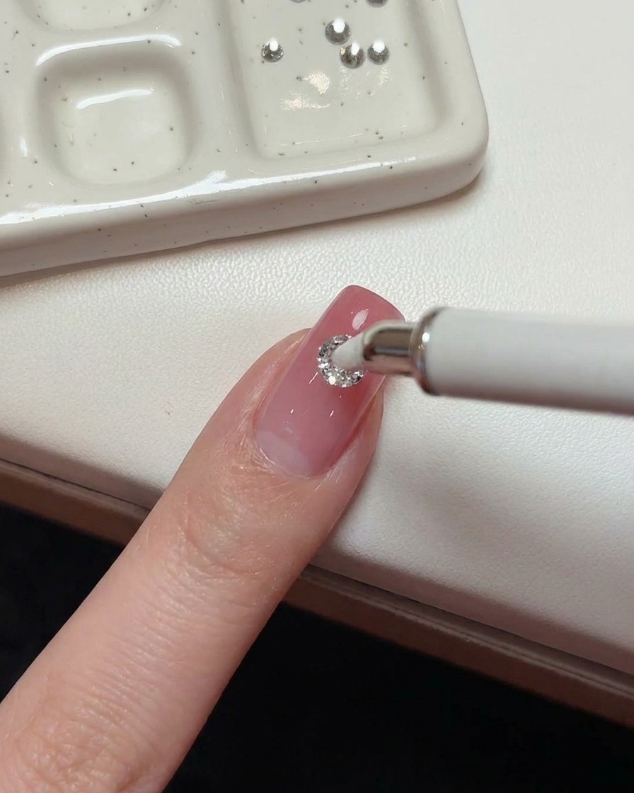 Un stylo blanc à pointe de cire plaçant un grand cristal rond de 5 mm au centre dun ongle rose.