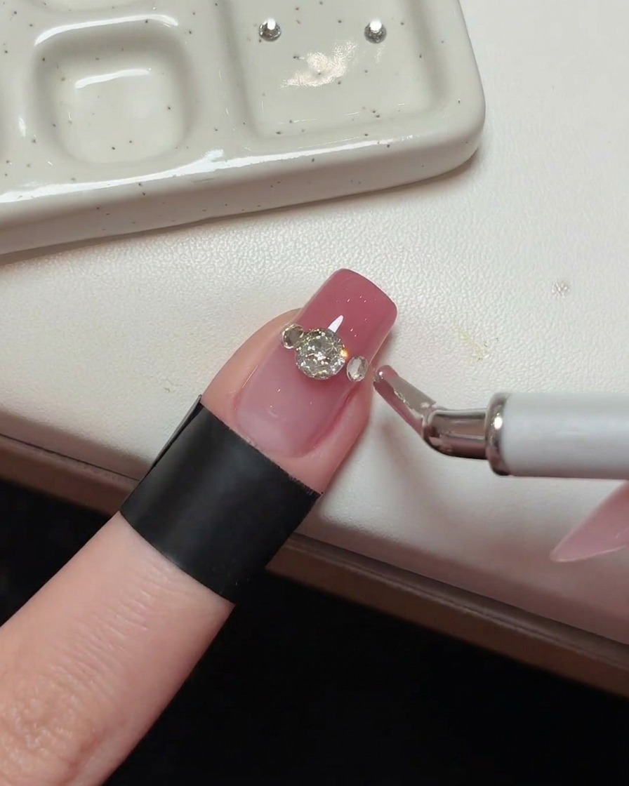 Gros plan dun cristal principal sur un ongle rose, avec des cristaux daccent en cours de placement sur les côtés.