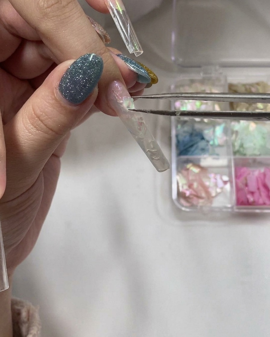 Une pince déposant de fines parcelles de coquillage irisé sur une couche de gel humide.