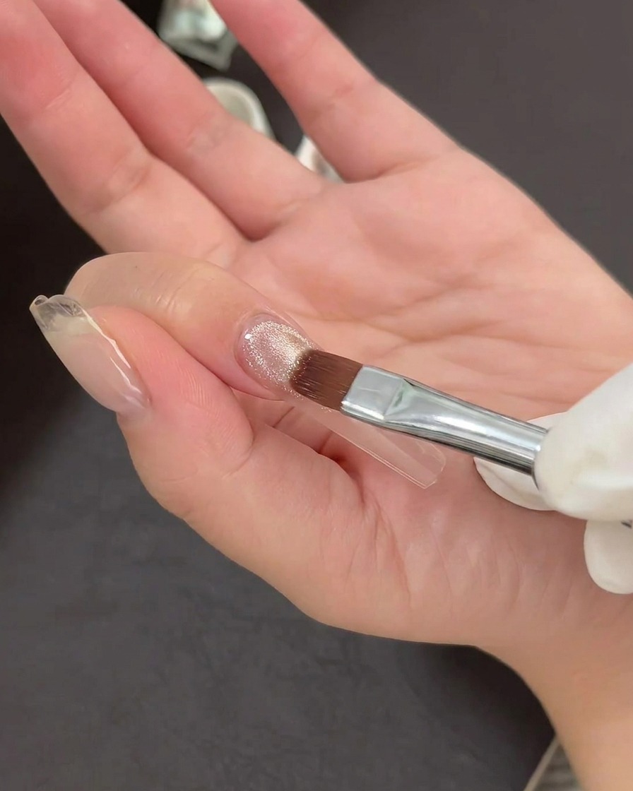 Un pinceau à nail art appliquant une fine couche de vernis gel cat-eye scintillant sur une pointe dongle transparente.
