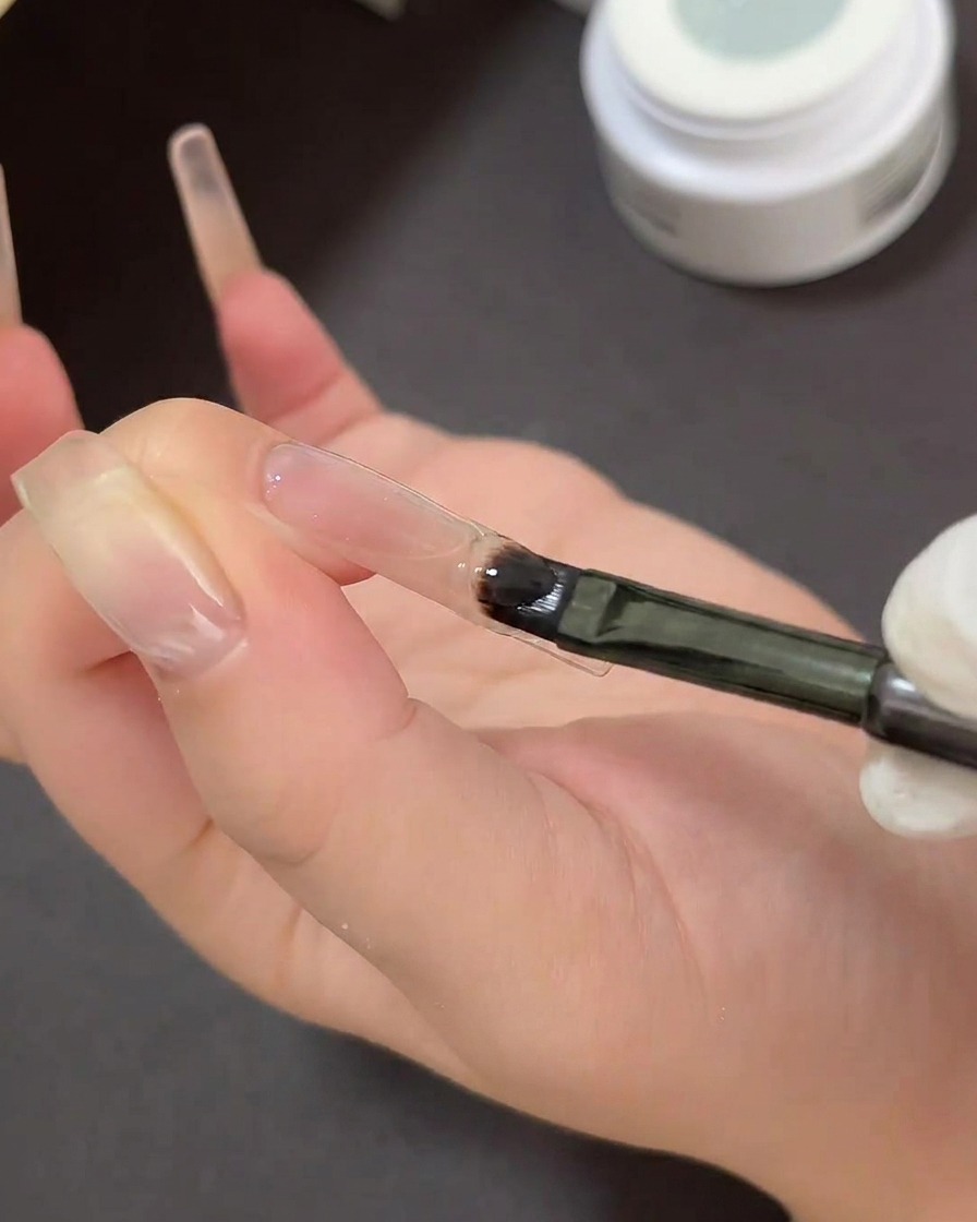 Un pinceau gel plat peignant du gel collant transparent sur la pointe dun ongle transparent au-dessus dun pochoir de French manucure incurvé.