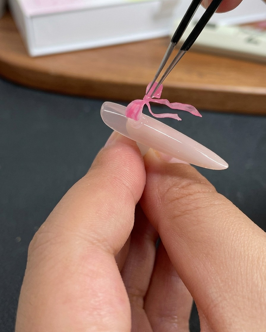 Une pince déposant un délicat charm papillon rose sur un ongle recouvert de gel transparent.