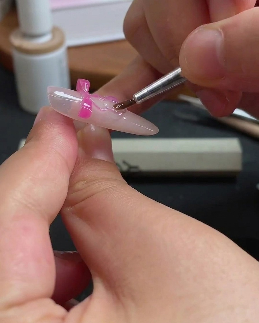 Un fin pinceau à ongles appliquant soigneusement du gel transparent autour des bords dun charm papillon rose sur un ongle.