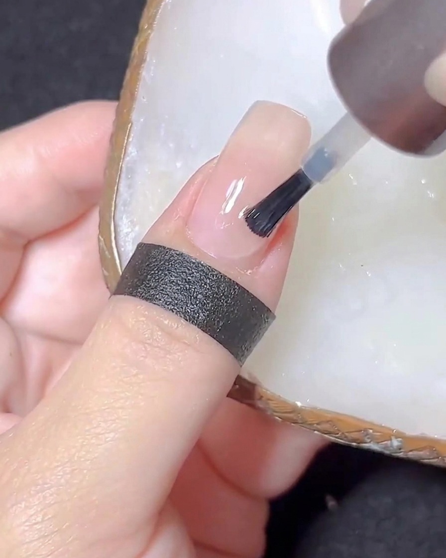 Un pinceau à ongles appliquant un top coat transparent sur un ongle de couleur nude.