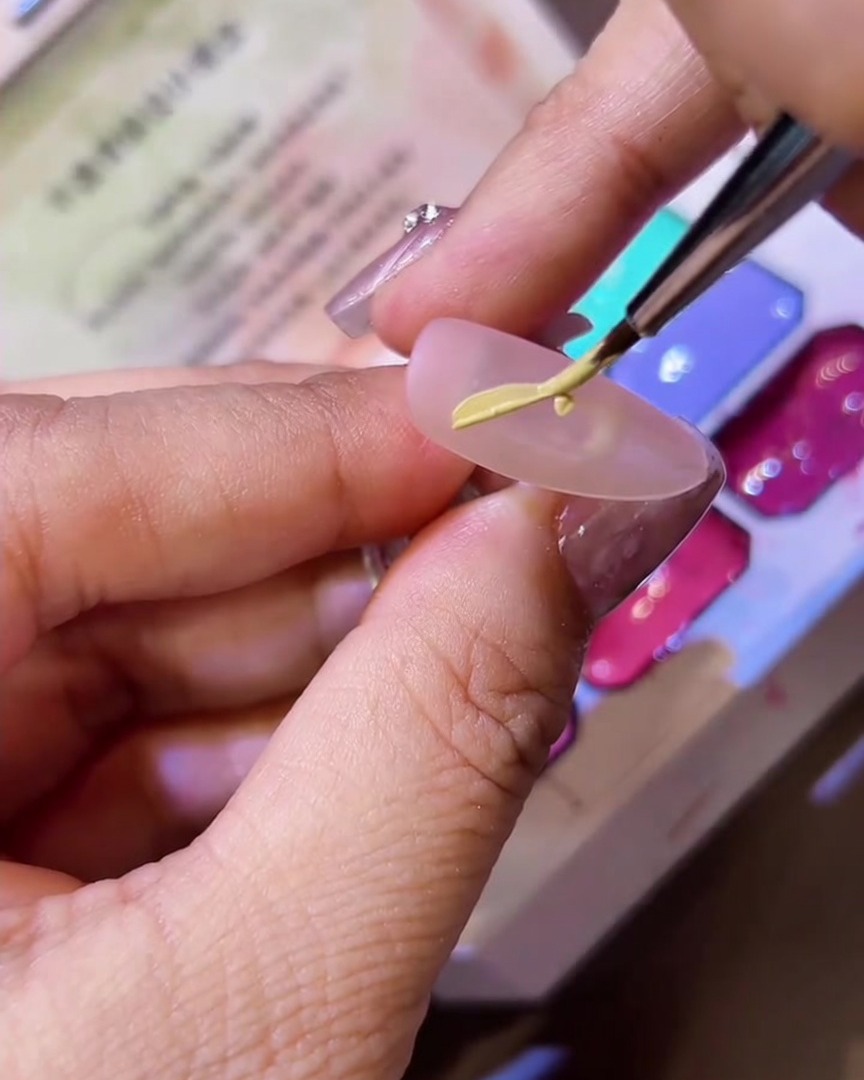 Un pinceau fin dessinant la forme dune demi-aile de papillon sur un ongle mat avec du vernis gel jaune.