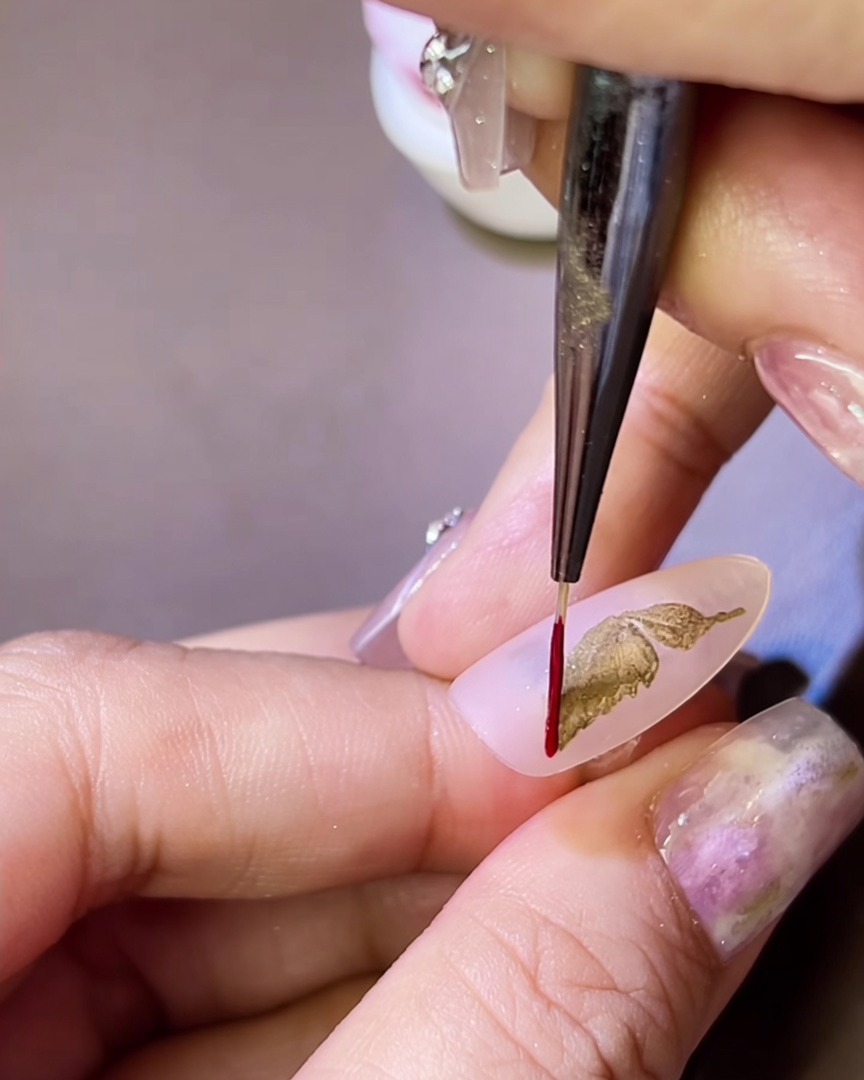 Un pinceau fin appliquant un fin contour en gel rouge autour dune aile de papillon métallique dorée sur un ongle.